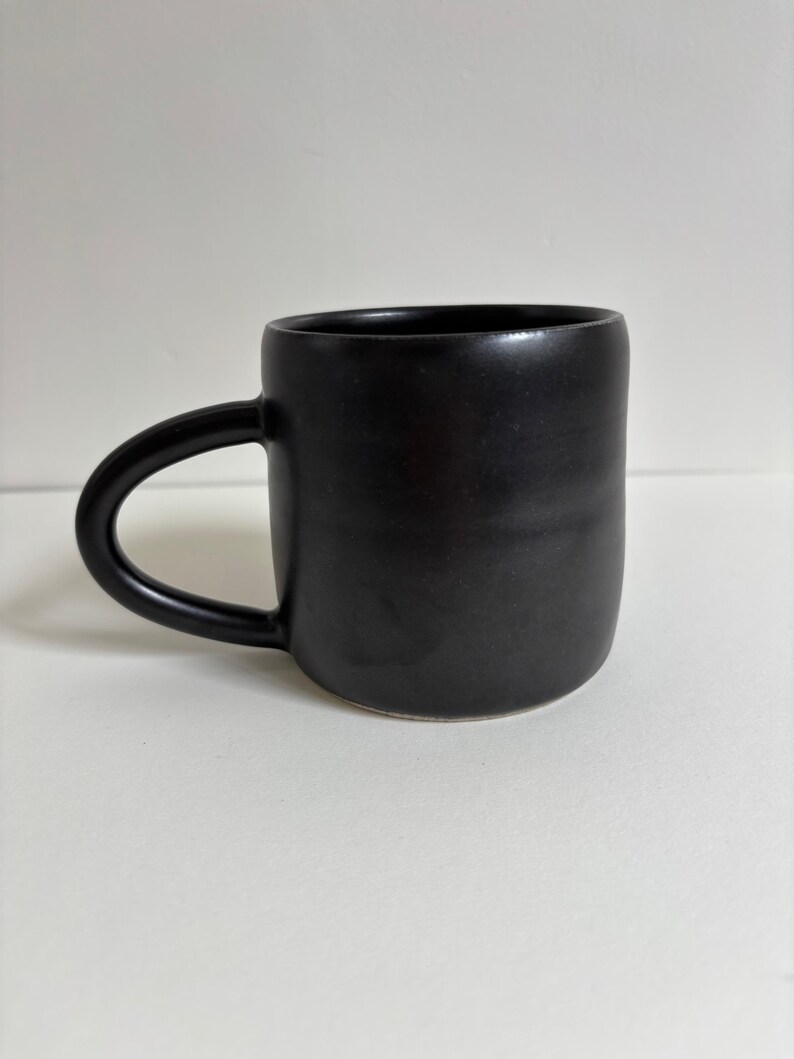 May include: A matte black ceramic mug with a smooth, cylindrical body and a curved handle on the left side. The mug features a solid, dark finish with subtle horizontal indentations along the exterior surface, creating a simple, uniform appearance.