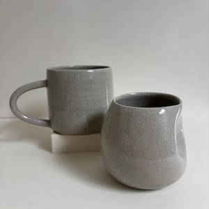 May include: Two light gray ceramic mugs with a speckled finish. One mug features a traditional curved handle, while the other is a rounded, handleless vessel with a thumb indentation on the side. Both items have a smooth, glazed surface.