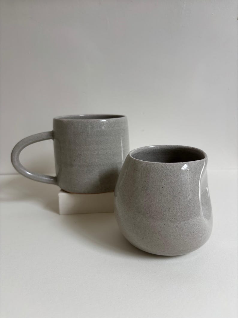 May include: Two light gray ceramic mugs with a speckled finish. One mug features a traditional curved handle, while the other is a rounded, handleless vessel with a thumb indentation on the side. Both items have a smooth, glazed surface.