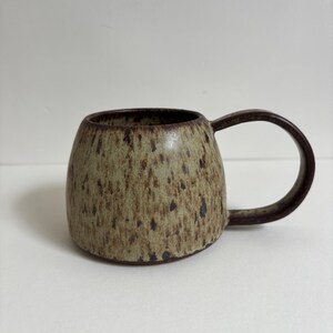 May include: A ceramic mug with a rounded body and a curved handle. The exterior features a speckled, light tan and brown glaze with a darker brown rim and handle. The surface has a textured appearance with visible dark brown spots throughout.
