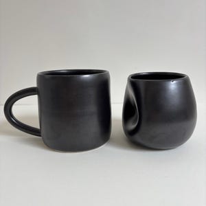 May include: Two matte black ceramic vessels. One is a cylindrical mug with a curved handle on the side. The other is a rounded, bowl-shaped cup featuring a distinct, concave indentation on one side, giving it a soft, irregular, and organic appearance.