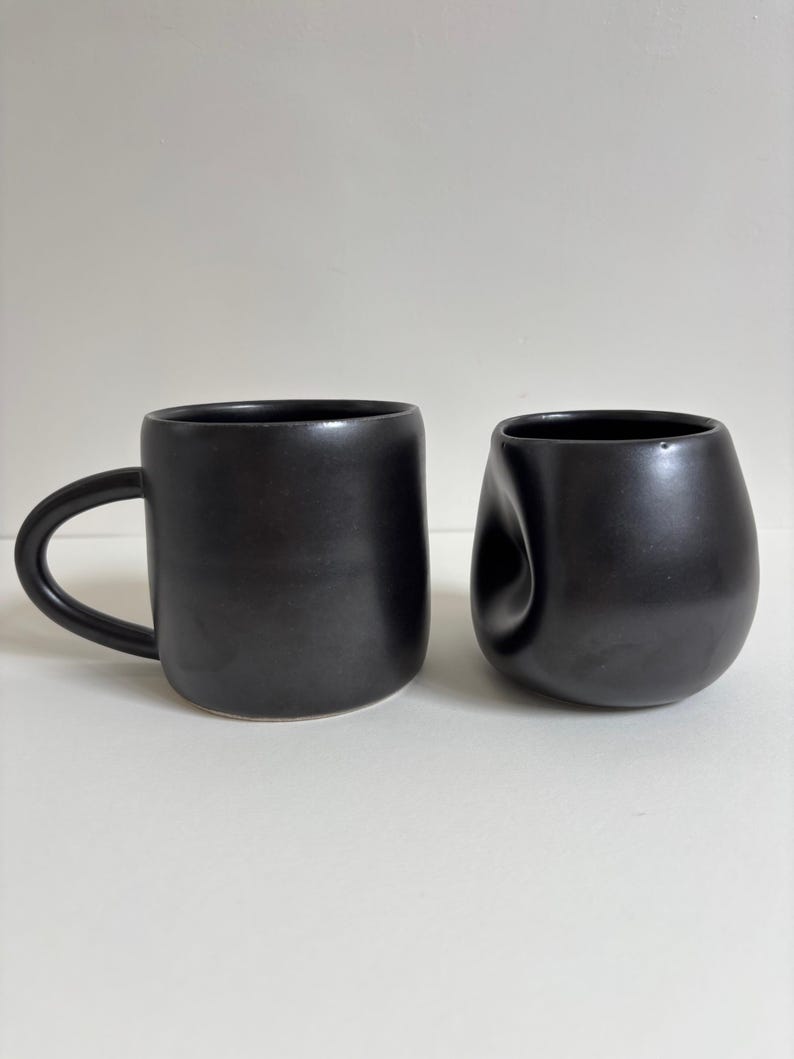 May include: Two matte black ceramic vessels. One is a cylindrical mug with a curved handle on the side. The other is a rounded, bowl-shaped cup featuring a distinct, concave indentation on one side, giving it a soft, irregular, and organic appearance.