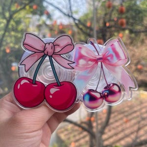 May include: Two acrylic charms featuring cherry pairs with bows. The left charm has solid red cherries and a light pink bow. The right charm features iridescent, metallic-toned cherries and a translucent, holographic-effect pink bow.
