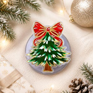 May include: A circular phone grip featuring a decorative acrylic topper shaped like a Christmas tree. The tree is dark green with white highlights, a brown trunk, and a large red bow at the top, all outlined with a shimmering gold glitter border.