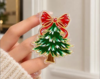 May include: A clear acrylic pin featuring a stylized green Christmas tree with white highlights and a brown trunk. The tree is topped with a large, vibrant red bow with gold glitter accents. The design is layered on a transparent acrylic base.