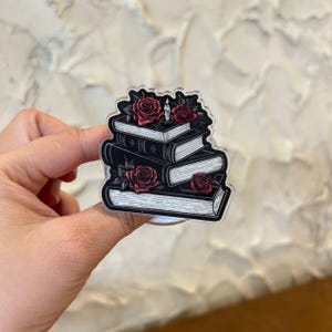 May include: An acrylic pin featuring a stack of three black and white books adorned with deep red roses and a small lit candle on top. The illustration is rendered in a detailed line art style with a clear acrylic border surrounding the design.