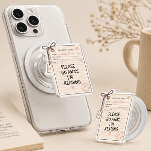 May include: A clear smartphone case with a collapsible phone grip featuring a rectangular library card design. The card is cream-colored with black lines and text that reads, "LIBRARY CARD," "book lover," and "PLEASE GO AWAY, I'M READING."