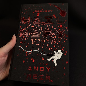 May include: A black hardcover book titled Project Hail Mary by Andy Weir. The cover features metallic red text and scattered red splatter patterns against a black background, with a white illustration of an astronaut tethered by a thin line.