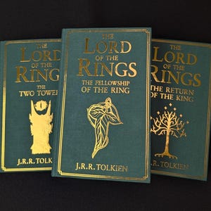 May include: Three hardback books titled The Lord of the Rings by J.R.R. Tolkien. The books feature dark teal covers with gold foil lettering and illustrations. The titles are The Two Towers, The Fellowship of the Ring, and The Return of the King.