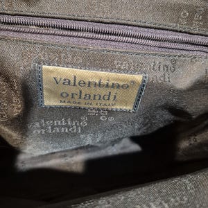 Pu&ograve; includere: Primo piano dell'interno di una borsa marrone scuro. Un'etichetta rettangolare recita "valentino orlandi MADE IN ITALY". La fodera della borsa &egrave; di una tonalit&agrave; di marrone pi&ugrave; scura con un motivo sottile. Una cerniera &egrave; visibile in alto.