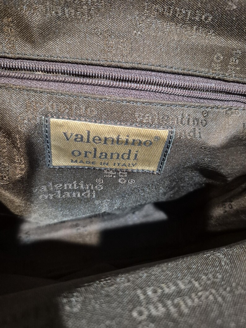 Pu&ograve; includere: Primo piano dell'interno di una borsa marrone scuro. Un'etichetta rettangolare recita "valentino orlandi MADE IN ITALY". La fodera della borsa &egrave; di una tonalit&agrave; di marrone pi&ugrave; scura con un motivo sottile. Una cerniera &egrave; visibile in alto.