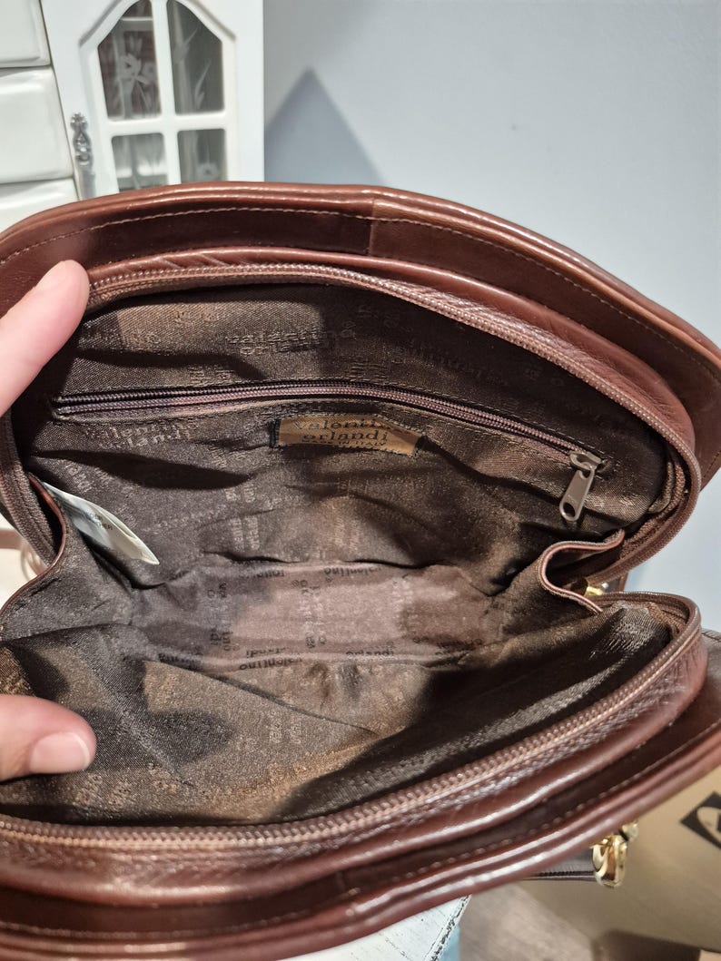 Pu&ograve; includere: Una borsa a mano in pelle marrone con chiusura a zip. L'interno &egrave; foderato con un tessuto fantasia. La borsa ha una forma arrotondata e una chiusura color oro. Il marchio "Valentino Orlandi" &egrave; visibile all'interno della borsa.