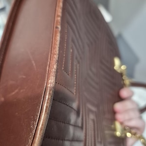 Pu&ograve; includere: Borsa a mano in pelle marrone con motivo trapuntato geometrico. La borsa ha una tracolla a catena dorata e una forma arrotondata. I bordi della borsa mostrano usura. La borsa &egrave; tenuta da una persona.