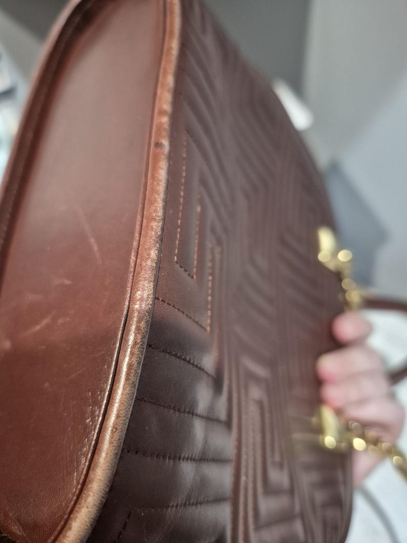 Pu&ograve; includere: Borsa a mano in pelle marrone con motivo trapuntato geometrico. La borsa ha una tracolla a catena dorata e una forma arrotondata. I bordi della borsa mostrano usura. La borsa &egrave; tenuta da una persona.
