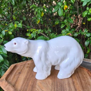 May include: A white ceramic polar bear figurine in a walking pose. The bear has a smooth, glossy finish, a black nose, and small dark eyes. The sculpture is depicted in profile, showing the textured details of the fur along the legs and body.