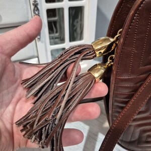 Pu&ograve; includere: Una borsa a tracolla in pelle marrone con un motivo geometrico e una lunga tracolla. La borsa presenta due nappe decorative con dettagli metallici color oro. Le nappe sono realizzate con sottili strisce di pelle.