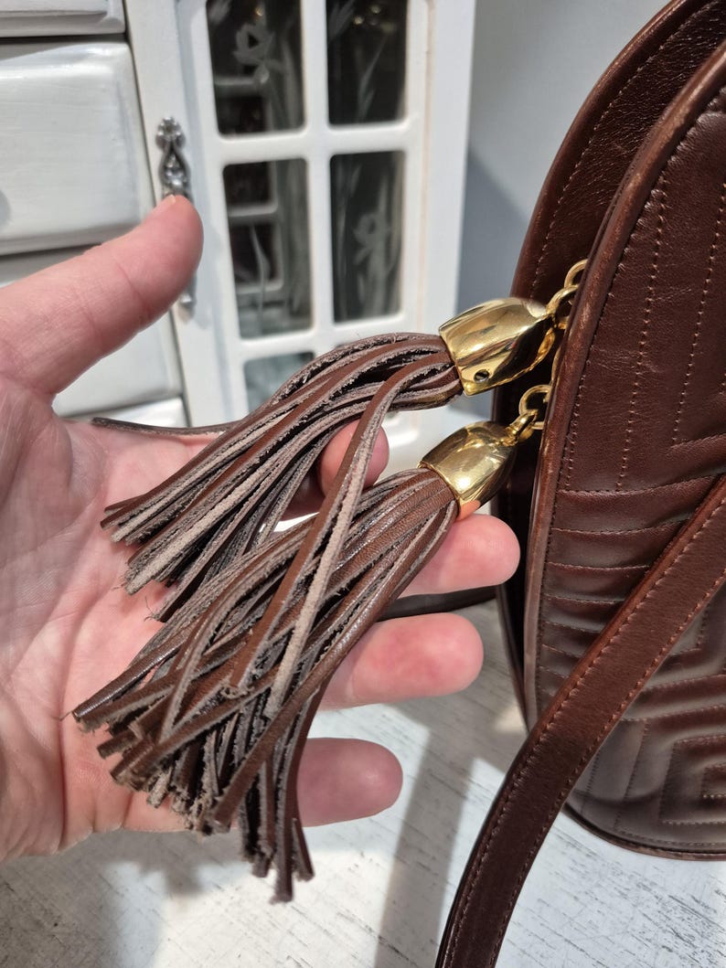 Pu&ograve; includere: Una borsa a tracolla in pelle marrone con un motivo geometrico e una lunga tracolla. La borsa presenta due nappe decorative con dettagli metallici color oro. Le nappe sono realizzate con sottili strisce di pelle.