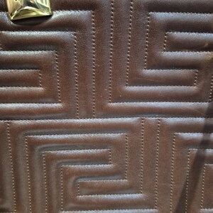 Pu&ograve; includere: Primo piano di una borsa in pelle marrone con un motivo geometrico. La borsa presenta un design cucito e un'etichetta metallica dorata con la scritta "valentino orlandi". La pelle ha un aspetto ricco e strutturato.