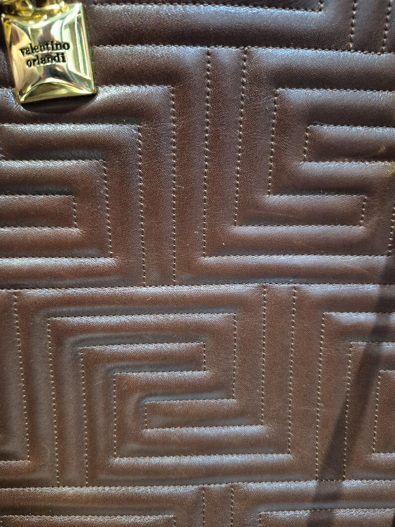 Pu&ograve; includere: Primo piano di una borsa in pelle marrone con un motivo geometrico. La borsa presenta un design cucito e un'etichetta metallica dorata con la scritta "valentino orlandi". La pelle ha un aspetto ricco e strutturato.
