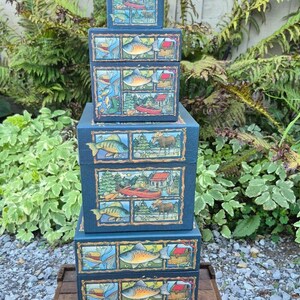 May include: A stack of four square dark blue boxes, each featuring a repeating decorative panel design. The illustrations on the boxes depict outdoor scenes including fish, a red canoe, a cabin, a moose, and fishing gear in a grid layout.