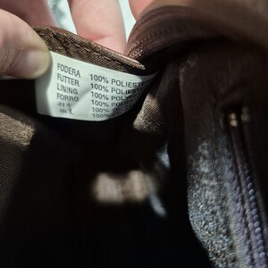 Pu&ograve; includere: Primo piano dell'interno di una borsa in tessuto marrone con un'etichetta. L'etichetta contiene testo in pi&ugrave; lingue, tra cui "FODERA" e "100% POLIESTER". L'interno della borsa &egrave; marrone scuro e la cerniera &egrave; visibile.