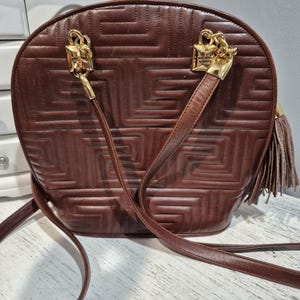 Pu&ograve; includere: Borsa a tracolla in pelle marrone con motivo geometrico. La borsa presenta elementi metallici color oro, tra cui due chiusure decorative e un nappa. Ha una lunga tracolla per essere indossata a spalla o a tracolla.
