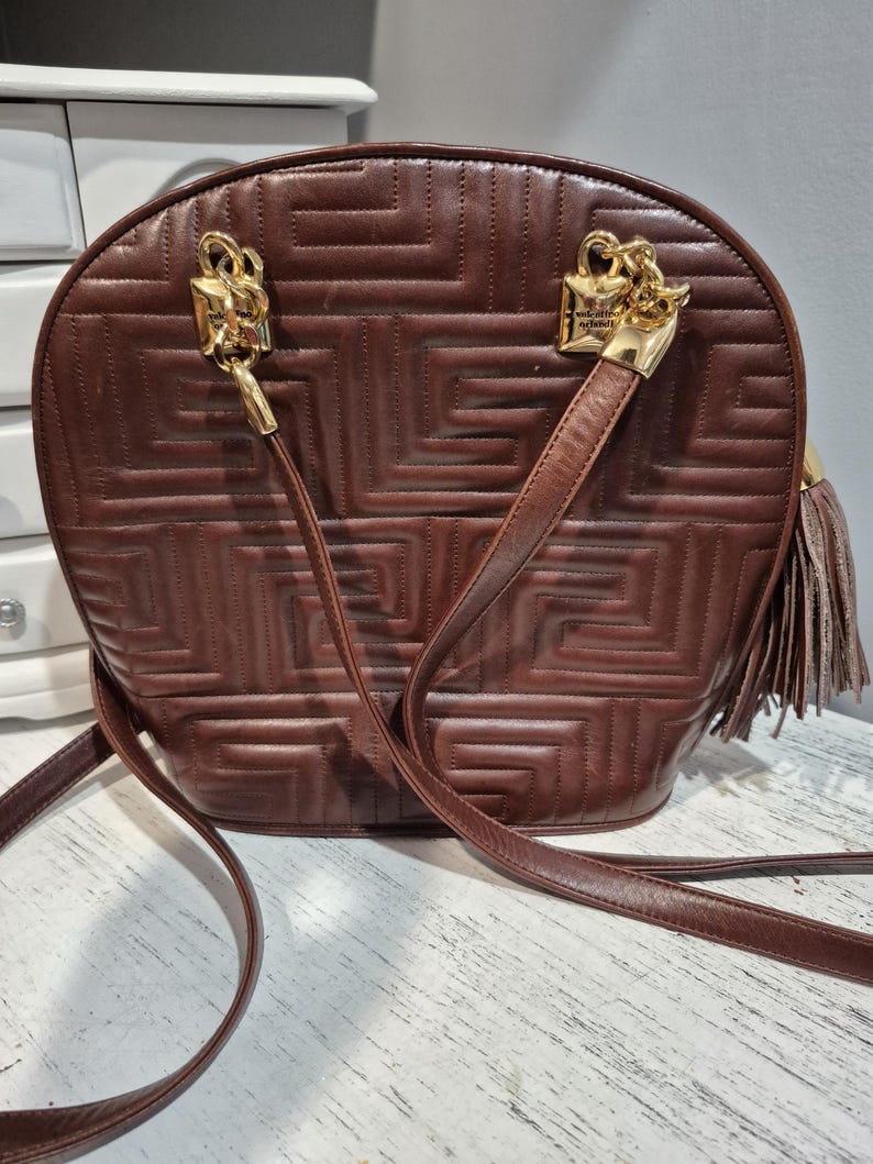 Pu&ograve; includere: Borsa a tracolla in pelle marrone con motivo geometrico. La borsa presenta elementi metallici color oro, tra cui due chiusure decorative e un nappa. Ha una lunga tracolla per essere indossata a spalla o a tracolla.
