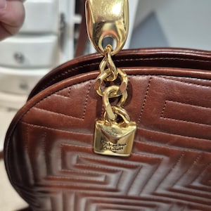 Pu&ograve; includere: Borsa a mano in pelle marrone con motivo geometrico, catena e chiusura dorate. La borsa ha un cinturino in pelle marrone e una chiusura in metallo dorato con il nome del marchio "Valentino Garavani" inciso.