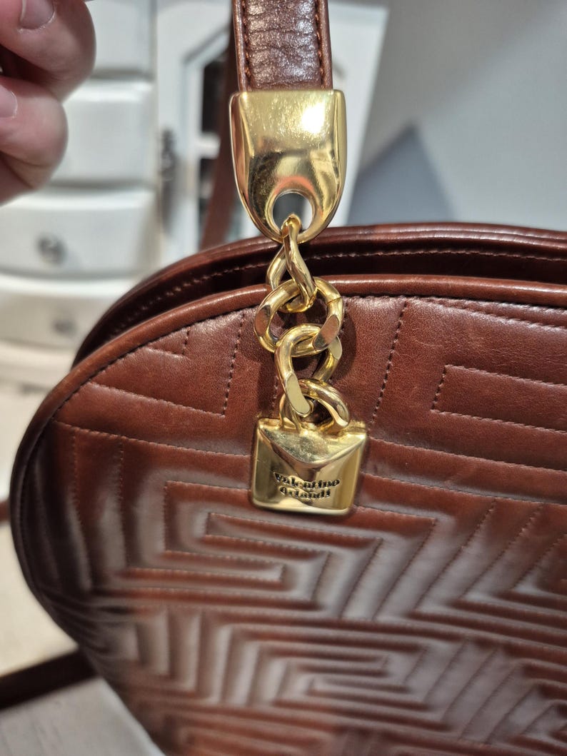 Pu&ograve; includere: Borsa a mano in pelle marrone con motivo geometrico, catena e chiusura dorate. La borsa ha un cinturino in pelle marrone e una chiusura in metallo dorato con il nome del marchio "Valentino Garavani" inciso.