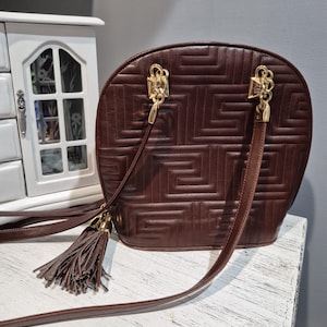 Pu&ograve; includere: Una borsa in pelle marrone con un motivo geometrico e finiture color oro. La borsa ha una lunga tracolla e un nappa. La borsa &egrave; appoggiata su una superficie bianca.