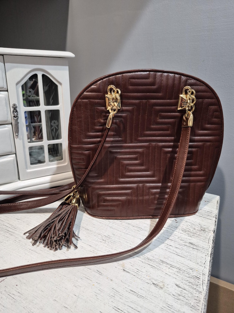 Pu&ograve; includere: Una borsa in pelle marrone con un motivo geometrico e finiture color oro. La borsa ha una lunga tracolla e un nappa. La borsa &egrave; appoggiata su una superficie bianca.