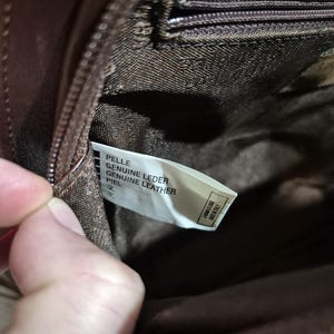 Pu&ograve; includere: Primo piano dell'interno di una borsa in pelle marrone. Un'etichetta bianca recita "PELLE GENUINE LEATHER PIEL" con testo in giapponese. La cerniera e la fodera della borsa sono visibili. L'etichetta dice anche "FABBRICATO IN ITALY".