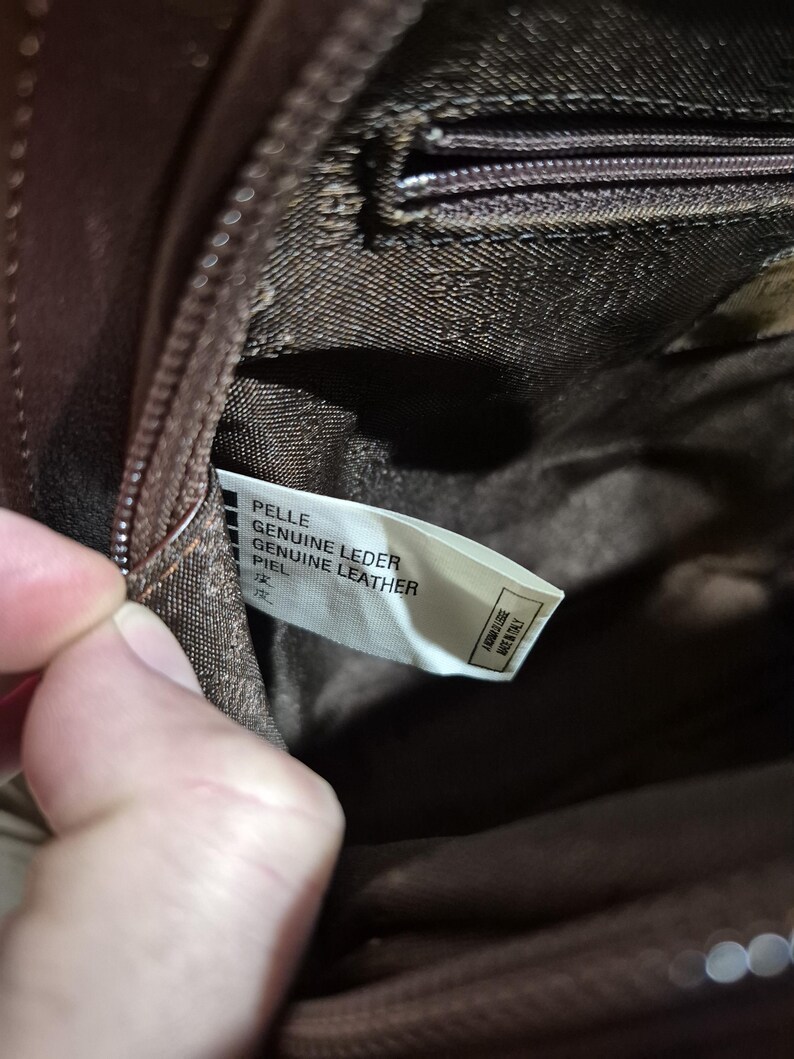 Pu&ograve; includere: Primo piano dell'interno di una borsa in pelle marrone. Un'etichetta bianca recita "PELLE GENUINE LEATHER PIEL" con testo in giapponese. La cerniera e la fodera della borsa sono visibili. L'etichetta dice anche "FABBRICATO IN ITALY".