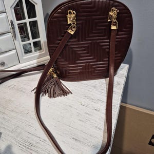 Pu&ograve; includere: Una borsa in pelle bordeaux con un motivo geometrico. La borsa ha finiture color oro, una lunga tracolla e un nappa decorativa. La borsa &egrave; su una superficie bianca.