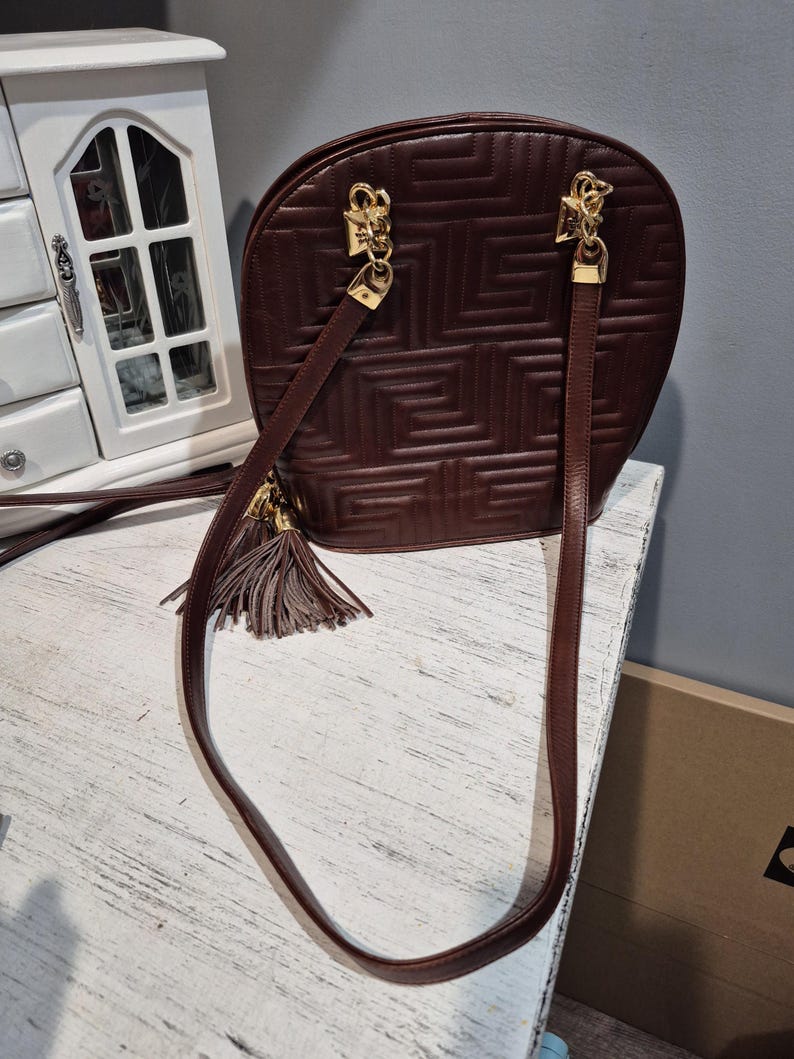 Pu&ograve; includere: Una borsa in pelle bordeaux con un motivo geometrico. La borsa ha finiture color oro, una lunga tracolla e un nappa decorativa. La borsa &egrave; su una superficie bianca.