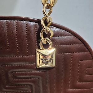 Pu&ograve; includere: Primo piano di una borsa in pelle marrone con manico a catena dorata. La borsa presenta un motivo geometrico e una chiusura quadrata dorata con la scritta "valentino orlandi".