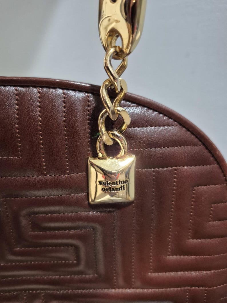 Pu&ograve; includere: Primo piano di una borsa in pelle marrone con manico a catena dorata. La borsa presenta un motivo geometrico e una chiusura quadrata dorata con la scritta "valentino orlandi".