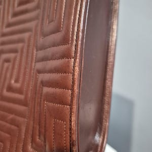 Pu&ograve; includere: Primo piano di una borsa in pelle marrone con un motivo trapuntato geometrico. La borsa ha un design strutturato con cuciture visibili e un bordo liscio e curvo. La pelle sembra avere una leggera lucentezza.