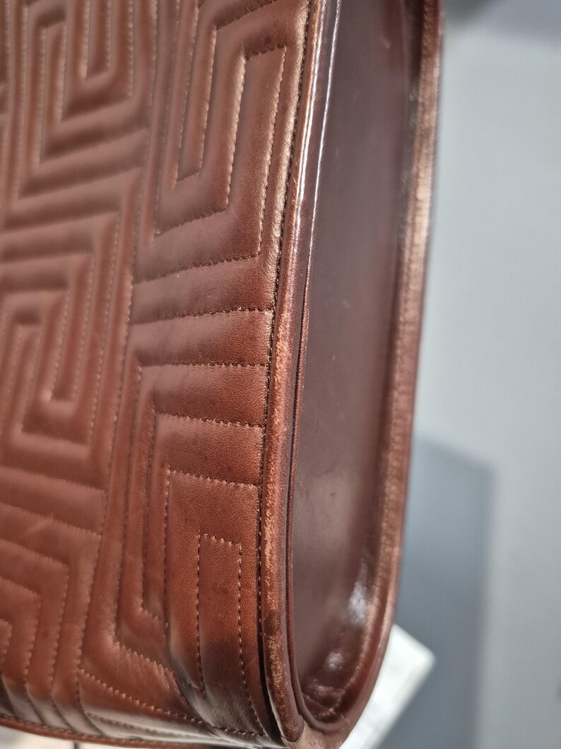 Pu&ograve; includere: Primo piano di una borsa in pelle marrone con un motivo trapuntato geometrico. La borsa ha un design strutturato con cuciture visibili e un bordo liscio e curvo. La pelle sembra avere una leggera lucentezza.