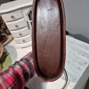 Pu&ograve; includere: Una borsa in pelle marrone con una lunga tracolla. La base della borsa &egrave; rettangolare con angoli arrotondati. La pelle ha un aspetto leggermente usurato.