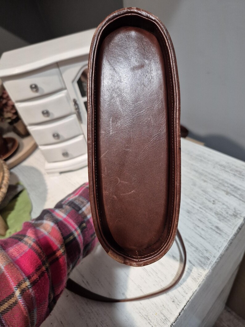 Pu&ograve; includere: Una borsa in pelle marrone con una lunga tracolla. La base della borsa &egrave; rettangolare con angoli arrotondati. La pelle ha un aspetto leggermente usurato.