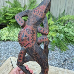 May include: A hand-carved wooden lizard sculpture featuring a dark brown finish with reddish-brown accents. The surface is decorated with black geometric patterns and a central circular motif containing a stylised animal figure on the lizard's belly.