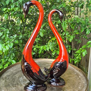 May include: Two ceramic swan figurines featuring a glossy finish with a striking color gradient. The base and lower bodies are a deep, dark brown, transitioning into a vibrant, bright orange that extends up the long, curved necks to the heads.