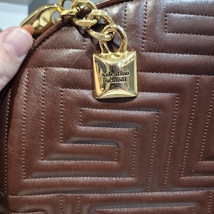 Pu&ograve; includere: Primo piano di una borsa in pelle marrone con un motivo geometrico. Una catena color oro e una chiusura rettangolare color oro con la scritta "valentino orlandi" sono attaccate alla borsa.