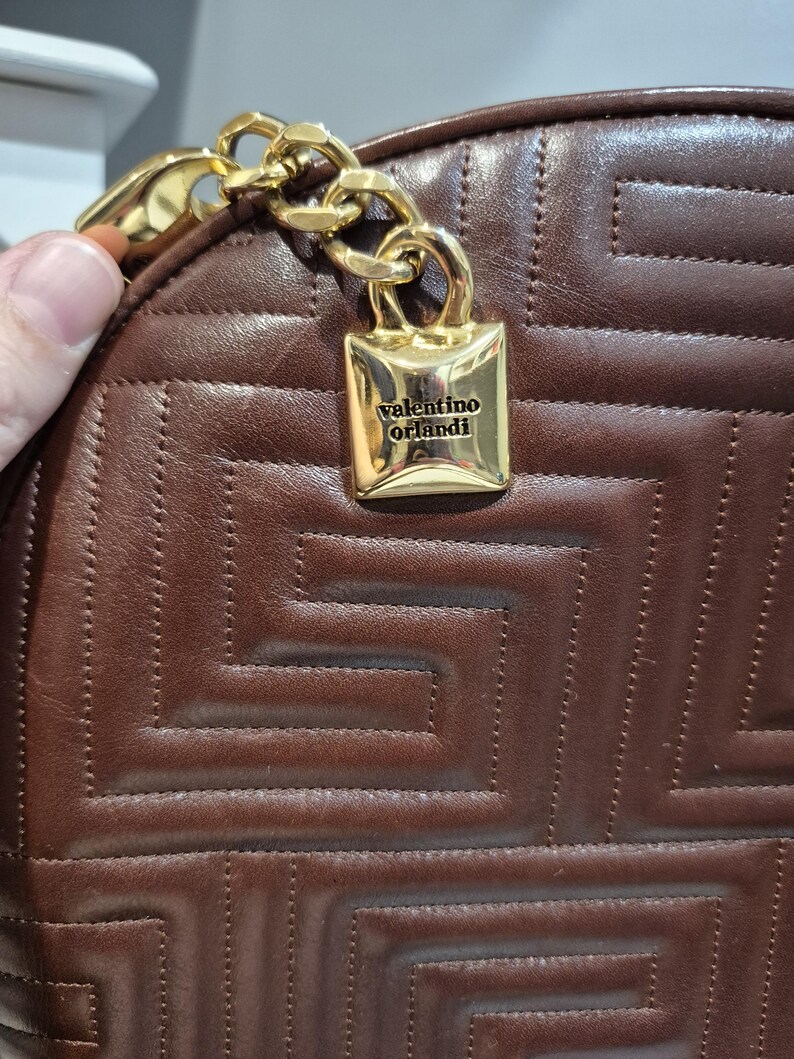 Pu&ograve; includere: Primo piano di una borsa in pelle marrone con un motivo geometrico. Una catena color oro e una chiusura rettangolare color oro con la scritta "valentino orlandi" sono attaccate alla borsa.