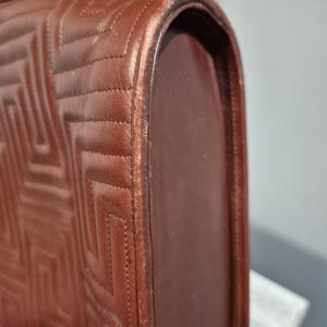Pu&ograve; includere: Primo piano di una borsa in pelle marrone con un motivo trapuntato geometrico. I bordi arrotondati della borsa mostrano segni di usura. Le cuciture sono visibili e la pelle ha un tono caldo.