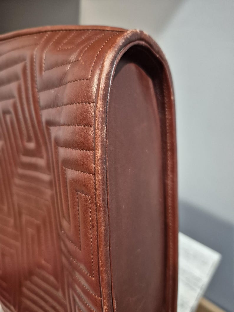Pu&ograve; includere: Primo piano di una borsa in pelle marrone con un motivo trapuntato geometrico. I bordi arrotondati della borsa mostrano segni di usura. Le cuciture sono visibili e la pelle ha un tono caldo.