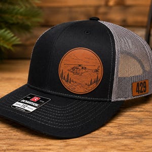 Puede incluir: Una gorra de camionero negra y gris con un parche circular de cuero marrón grabado con un helicóptero sobre un paisaje montañoso. Un pequeño parche rectangular de cuero marrón con el número 429 está fijado en el lateral de la malla trasera.