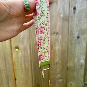 May include: A floral patterned lanyard with a pink and green design. The lanyard features a repeating pattern of pink roses and green leaves on a cream background. It has a metal clasp and key ring.