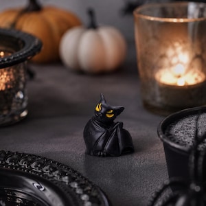 May include: A Halloween-themed still life featuring a black bat figurine with yellow eyes, pumpkins, a candle in a glass holder, and a decorative plate. The scene is set on a dark gray surface, creating a spooky atmosphere.